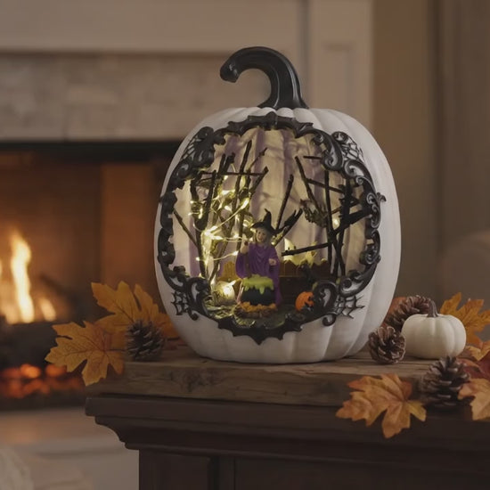 A slow cinematic zoom video of a lighted white resin pumpkin decoration glowing on a fireplace mantel at night, highlighting the intricate details of the internal miniature witch and bubbling cauldron scene.