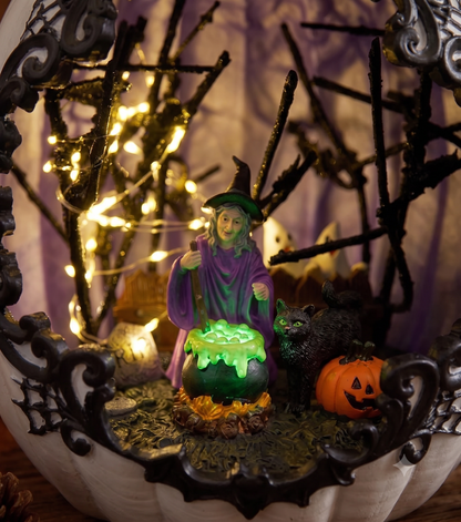 Macro close-up shot of the internal miniature scene showing a detailed witch figurine, a bubbling green cauldron, and a black cat illuminated by warm LED fairy lights